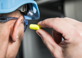 Employee wearing hard hat and protective eye glasses putting on ear plugs
