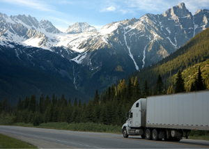 Semi truck driving down forest highway in front of snowy mountains