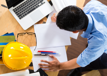 Birds-eye view of a business man leaning over a desk with papers, laptop, and a hard hat
